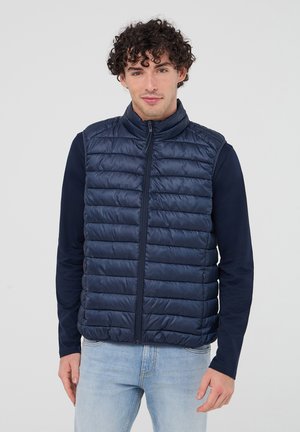 Gilet imbottito blu navy con colletto alto, chiusura con zip e texture a righe orizzontali. Indossato sopra una camicia blu navy a maniche lunghe e jeans chiari.
