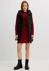 Chaleco acolchado negro con un diseño de rombos y capucha, superpuesto a un vestido rojo ceñido. La modelo lleva botas de combate negras con cordones.