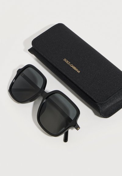 Μεγάλα τετράγωνα μαύρα γυαλιά ηλίου δίπλα σε μαύρη θήκη με το λογότυπο Dolce & Gabbana σε λευκό φόντο.