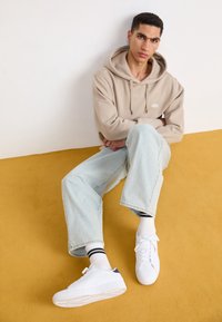 Sweat à capuche beige avec cordon, jean ample bleu clair, et baskets blanches avec des accents bleu marine. Le modèle est assis sur une surface de couleur moutarde.