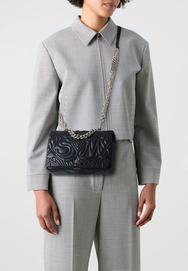 Sac à main en cuir matelassé noir avec un motif en relief ; comprend une bandoulière en chaîne et une fermeture magnétique. Associé à une chemise à carreaux.