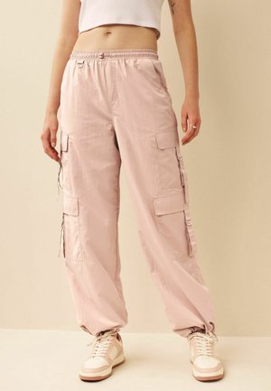 Person trägt hellrosa Cargo-Hosen mit mehreren Taschen, ein weißes, kurz geschnittenes Tanktop und beige Turnschuhe und steht vor einem neutralen Hintergrund.