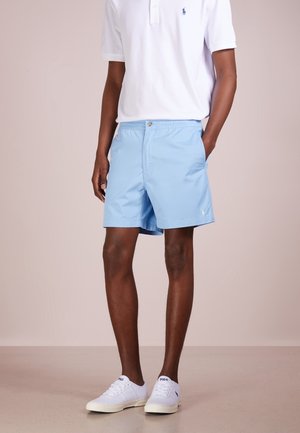 Homme portant un short bleu clair, un polo blanc et des baskets blanches, debout avec une main dans la poche sur fond uni.