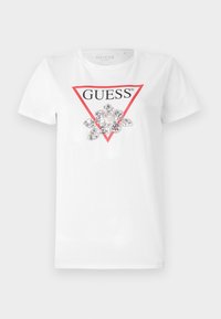 Balta marškinėliai su raudonu trikampiu logotipu, ant kurio užrašas „GUESS“, centre papuošti sidabriniais blizgučiais, kurie suformuoja gėlytės formą.