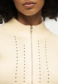 Κοντινό πλάνο ενός ατόμου που φοράει πλεκτό πουλόβερ σε εκρού χρώμα με ψηλό γιακά και ασημένιο φερμουάρ μπροστά.