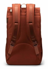 Sac à dos marron en tissu rembourré, avec des bretelles ajustables et une poignée supérieure. Il présente une texture lisse et une poche zippée sur le côté.