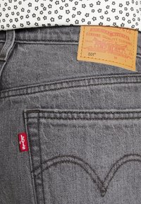 Grå denimjeans med en läderetikett som visar "Genuine Quality Clothing." Har röd Levi's-etikett och karakteristisk bakfickssömnad.