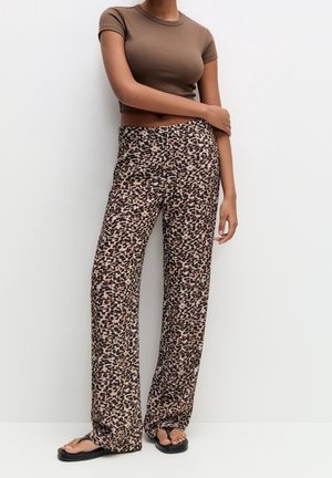 Vrouw draagt een bruin kort katoenen topje met korte mouwen, luipaardprint wijde broekspijpen en zwarte slippers, staand tegen een witte muur.