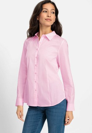 Woman looking sideways wearing a long-sleeve light pink buttoned shirt and blue jeans against a white background.