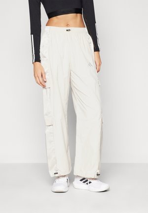 Persoon draagt witte losse cargobroek, witte Adidas sneakers en een zwart cropped top met lange mouwen, staand tegen een effen achtergrond.
