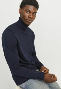 Morska pulover s turtleneck ovratnikom, izdelan iz mehkega materiala, z dolgimi rokavi in ribastimi manšetami. Model sedi ob nevtralni ozadju.