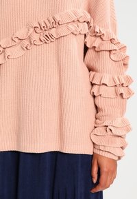 Jasnoróżowy dziergany sweter z falbankami na rękawach i przodzie, zestawiony z ciemnoniebieską plisowaną spódnicą.