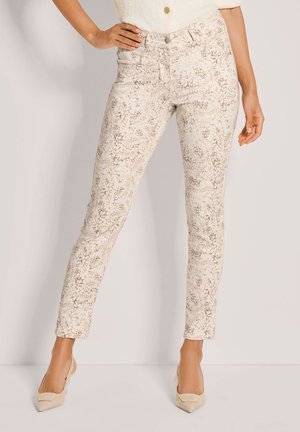 Femme portant un pantalon beige slim à motif léger serpent, associé à des talons pointus beige, debout avec une main sur la hanche.