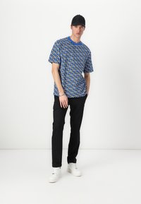 Blaues Grafik-T-Shirt mit einem sich wiederholenden Muster, kombiniert mit schwarzen Hosen und weißen Sneakers. Die Kappe ist schwarz und enganliegend.