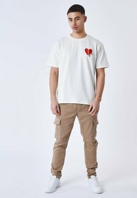 Maglietta bianca a maniche corte con una grafica di un cuore spezzato rosso sul lato sinistro del petto, abbinata a pantaloni cargo beige e scarpe da ginnastica bianche.