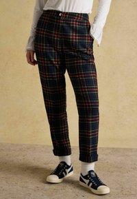 Raibās, augsta vidukļa bikses tumši zilā krāsā ar sarkaniem un zaļiem svītrām, kombinētas ar tumši zilām sporta kurpēm un baltām zeķēm, apšļautu biksēm.