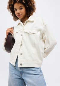 Νεαρή γυναίκα με κατσαρά μαλλιά, που φοράει λευκό τζιν μπουφάν και ανοιχτό μπλε τζιν παντελόνι, κρατώντας καφέ σκούρο δερμάτινη τσάντα στον ώμο.