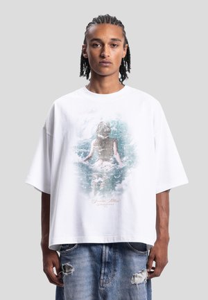 Jeune homme aux cheveux tressés portant un t-shirt blanc oversize avec une illustration d'une personne dans l'eau et un jean bleu déchiré.