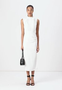 White sleeveless midi dress with gathered detail at the waist. Model holds a black handbag and wears black ankle-strap heels. Smooth fabric texture.