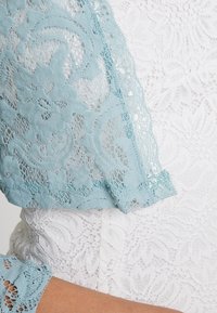 Kanten overlay in lichtblauw valt over witte bloemenkantstof, met complexe patronen en gegolfde randen. De textuur lijkt zacht en delicaat.