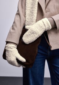 Helle beige Jacke mit einem strukturierten cremefarbenen Schal, kombiniert mit flauschigen weißen Fäustlingen. Hält eine braune Wildledertasche in der Hand zu Jeans aus Denim.