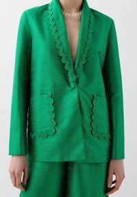 Green blazer in a textured fabric, featuring a scalloped neckline, front button, and pockets with scalloped edges. Styled with matching bottoms.