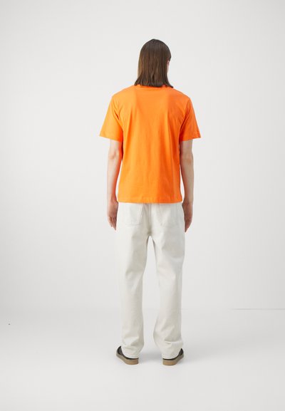 Orange cotton t-shirt with short sleeves, paired with cream-colored loose-fit jeans and black shoes. Seen from the back against a neutral background.