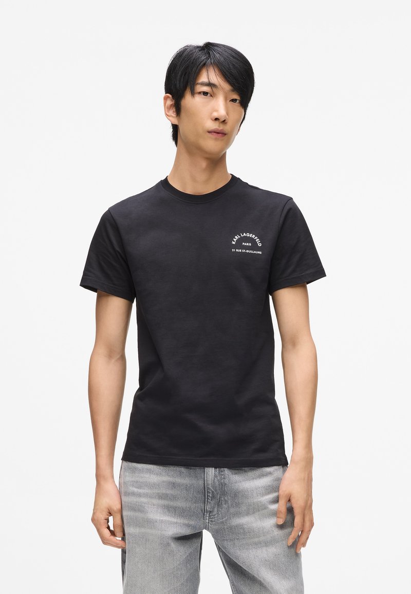 Camiseta de algodón negra con cuello redondo y mangas cortas. En la parte frontal tiene un texto impreso en blanco con un diseño curvado. Se lleva con unos jeans gris claro.