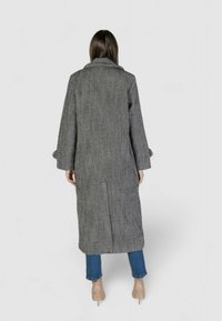 Cappotto lungo grigio e nero a motivo houndstooth con colletto a pistagna, maniche lunghe e spacchi laterali, abbinato a jeans blu e tacchi nudi.