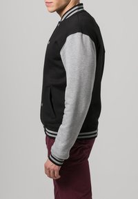 Zwart en grijs varsityjack met een zachte stof, gestreepte ribgebreide manchetten en zijzakken, gedragen over bordeauxrode broeken.