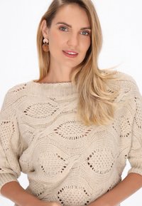 Pulover tricotat bej cu o textură în model de valuri, guler larg și mâneci până la trei sferturi. Cerceii au un design circular, bicolor.