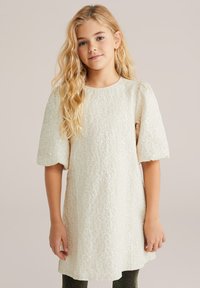 Čipkasta obleka v kremni barvi s kratkimi napihnjenimi rokavi, zaobljenim izrezom, teksturiranim cvetličnim vzorcem in ohlapnim krojem, v kombinaciji s temnimi hlačami.