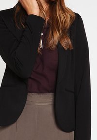 Blazer negro con un diseño ajustado, tejido texturizado y solapas con muesca, usado sobre una blusa de color púrpura oscuro, combinado con una falda marrón claro.