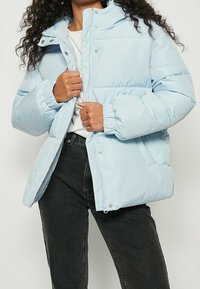 Lyseblå dunjakke med høj krave, trykknapper, elastiske manchetter og sidelommer. Materialet ser glat og isoleret ud.