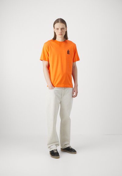 Short-sleeve orange cotton t-shirt with a black letter “É” on the left chest, paired with light-colored loose-fitting jeans and black sneakers.