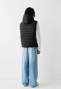 Czarna pikowana kamizelka z kapturem, z poziomym wzorem pikowania. Noszona na białej koszuli z długim rękawem, zestawiona z jasnoniebieskimi szerokimi spodniami jeansowymi.