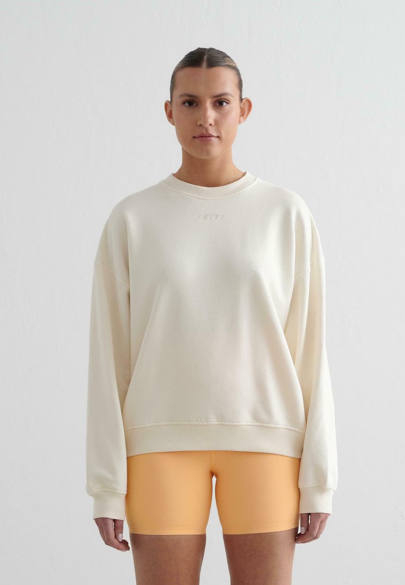 Felpa oversized color crema con polsini e orlo a coste, abbinata a pantaloni biker arancioni. Tessuto morbido, vestibilità casual, branding minimo.
