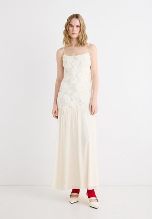 Frau trägt ein langes, weißes ärmelloses Kleid mit floraler Applikation am Oberteil und fließendem Rock, kombiniert mit roten Socken und weißen, spitzen Schuhen.