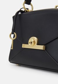 Borsa in pelle nera con chiusura a lucchetto dorato e una chiave dorata pendente attaccata al manico su uno sfondo bianco.