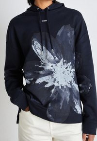 Mörkblå hoodie med stor grå blomgrafik, dragsnören och logotyp på bröstet. Mjuk tygkvalitet med en avslappnad passform och sidofickor.