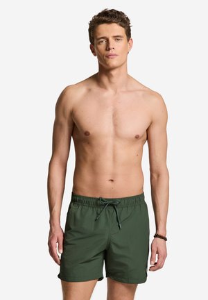 Nuori mies seisoo ilman paitaa päällään vihreät uimashortsit nyörillä, neutraali ilme ja rannekoru oikeassa ranteessa.