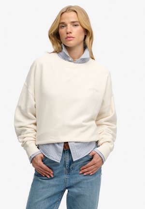 Blonde woman wearing a cream sweatshirt layered over a blue and white striped button-down shirt, with blue jeans, hands in pockets.
