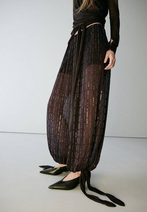Mujer que lleva pantalones anchos y translúcidos en negro con rayas verticales y puños atados, combinados con zapatos negros de punta puntiaguda y una blusa de manga larga.