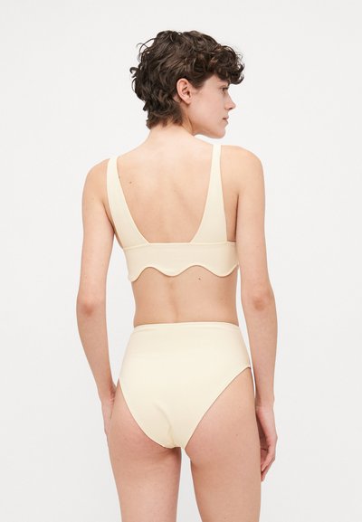 Krēmskrāsas bikini komplekts ar viļņotu malu uz topa, masīvām siksnām un augstām biksēm. Materiāls izskatās gluds un elastīgs.
