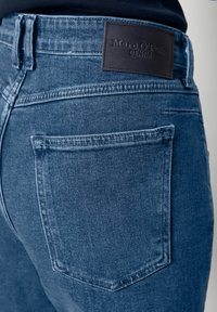 Blå denimjeans med en slät textur, med en brun läderlapp märkt "Marc O'Polo DENIM" på baksidan av midjebandet och en bakficka.
