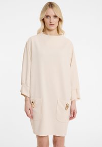 Beiges Kleid mit loser Silhouette, Dreiviertelärmel, zwei vorderen Taschen verziert mit metallischen Akzenten und einem runden Ausschnitt.