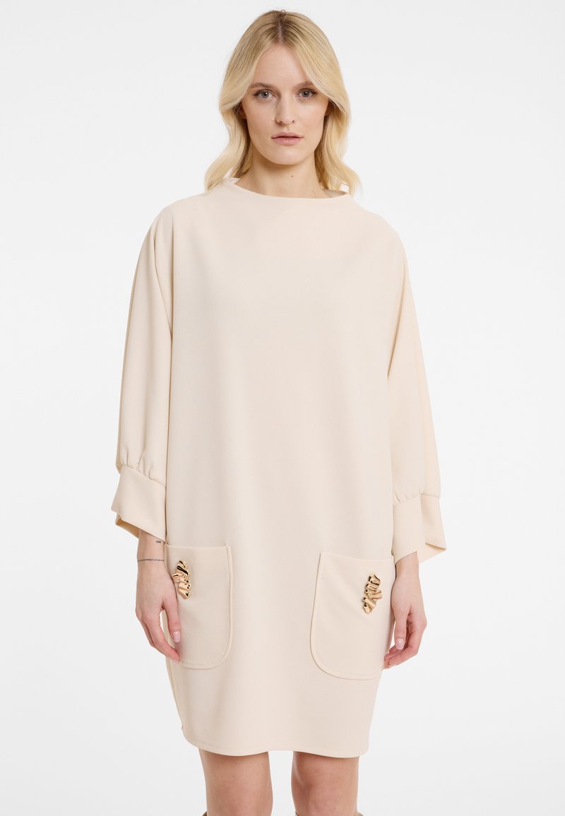 Beiges Kleid mit loser Silhouette, Dreiviertelärmel, zwei vorderen Taschen verziert mit metallischen Akzenten und einem runden Ausschnitt.