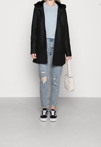 Abrigo negro con capucha sobre un suéter azul claro, combinado con jeans rasgados azul claro y zapatillas negras. Los accesorios incluyen un bolso tejido blanco.