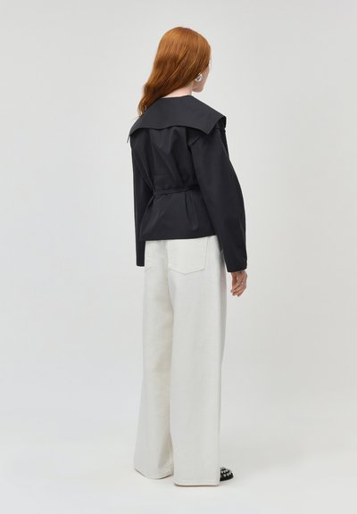 Woman with red hair wearing a black jacket with a wide collar and white wide-leg pants, standing against a plain light background.