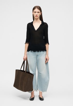 Woman stands holding large dark brown tote bag, wearing black long-sleeve top, light blue wide-leg jeans, and black flats on white background.